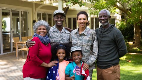 Portrait of a young adult African American male soldier and a young adult mixed race female soldier with thier diverse multi-generation family in the garden outside their home, embracing and smiling to camera, a US flag draped over the shoulders of the kids