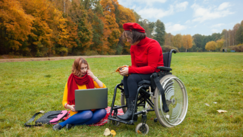 young adult female sitting on grassy lawn with laptop open. male in wheelchair beside her. autumn colored trees in background