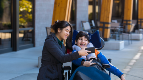 older sister holding phone for younger sibling in wheelchair. both smiling