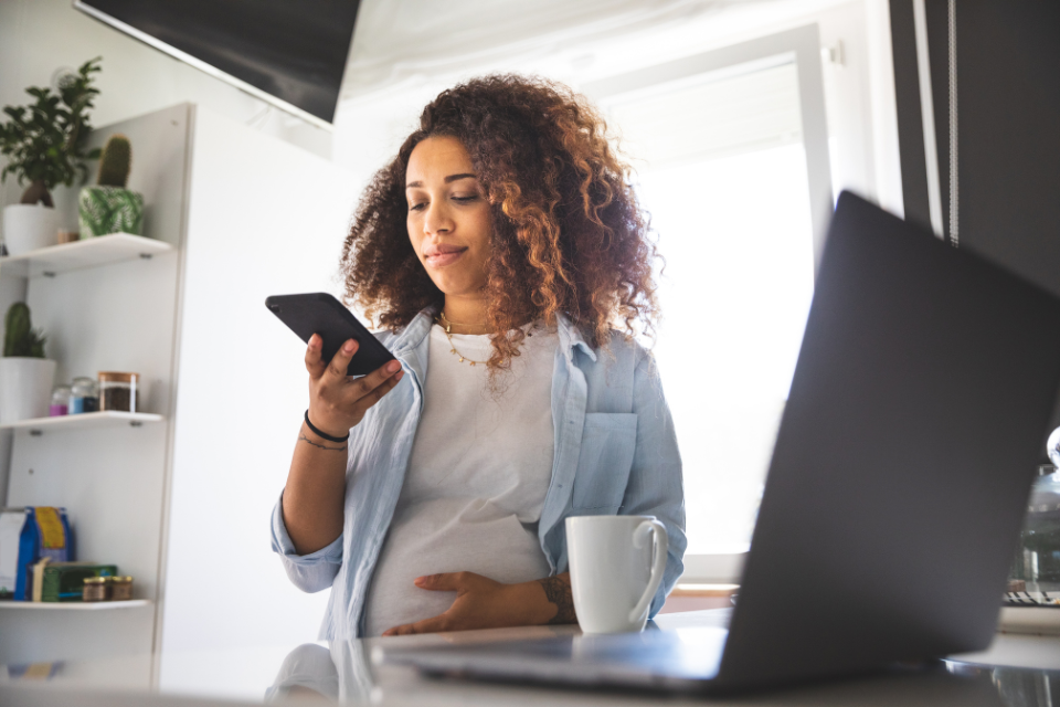 pregnant mom looking at phone while sitting in front of laptop