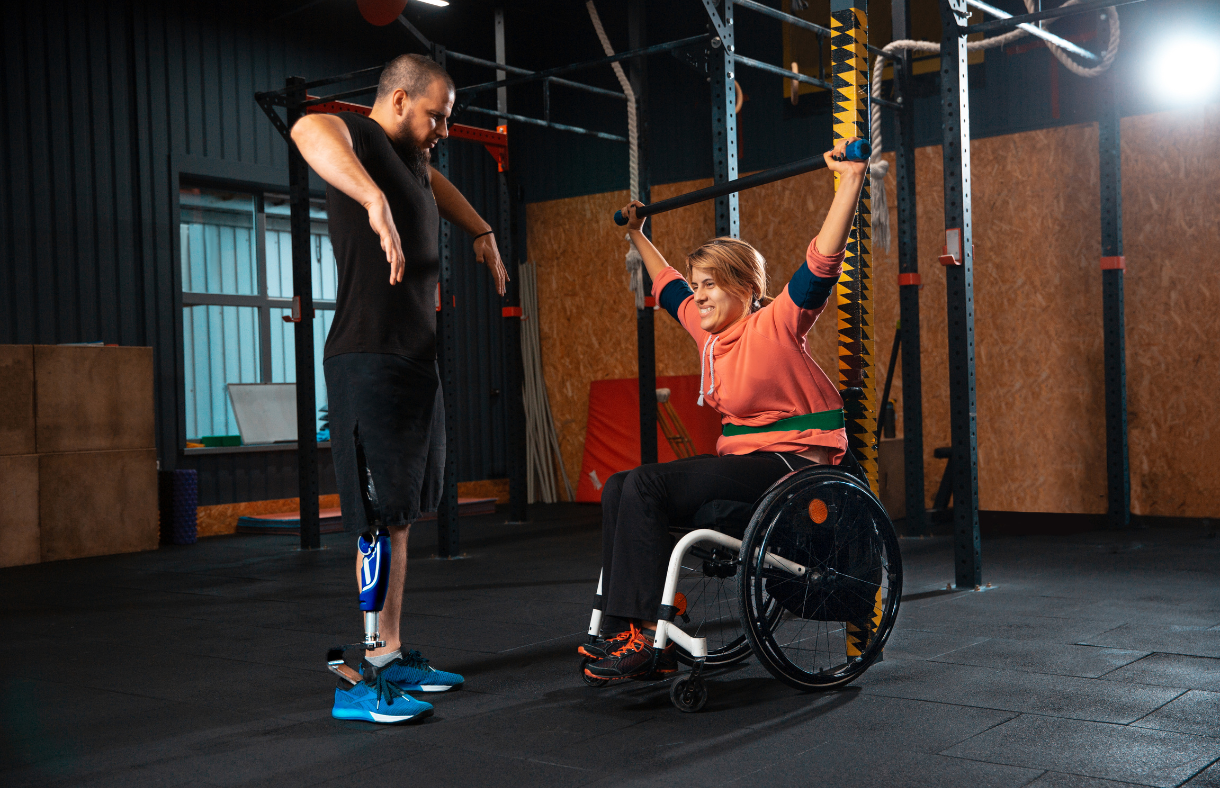 woman in wheelchair using pull bar in gym being assisted by trainer