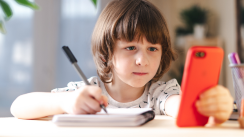 girl sitting at desk looking at phone while making a list with pencil and paper