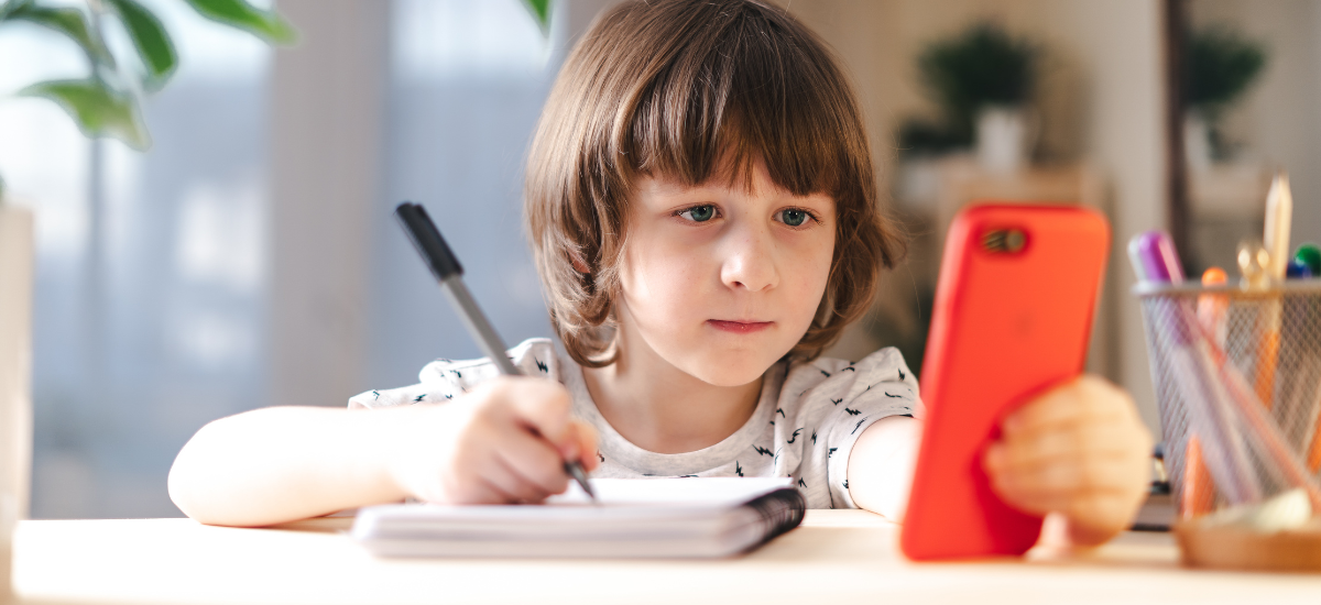 girl sitting at desk looking at phone while making a list with pencil and paper