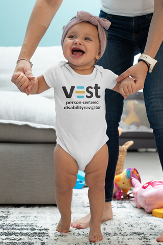 Baby in white Vest onesie, being supported in walking position by an adult standing behind and visible from the waist down.
