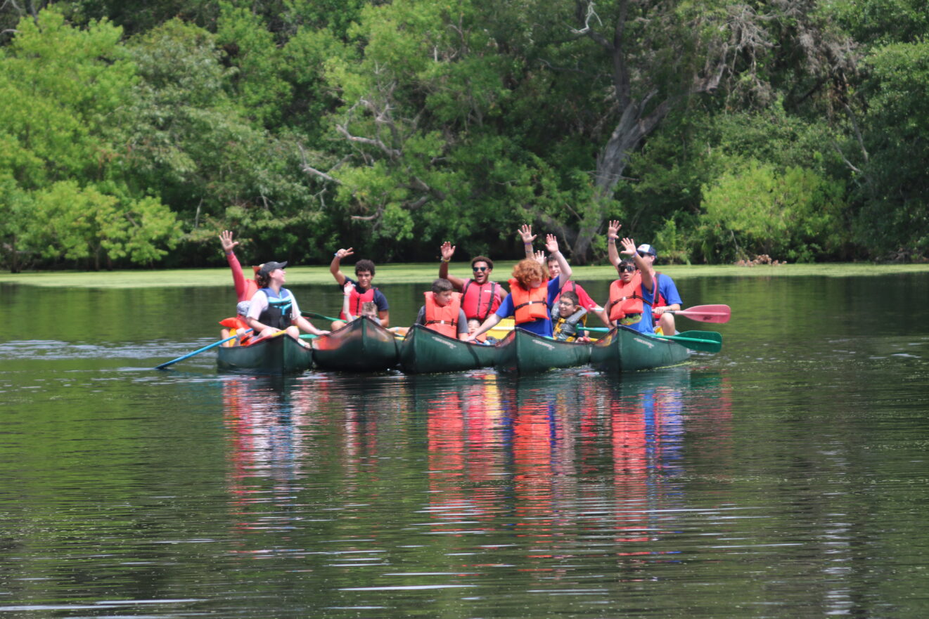 Ten ESGH campers waving from five green canoes on a lake