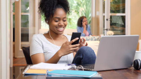 teen girl doing homework in front of laptop while also looking at phone