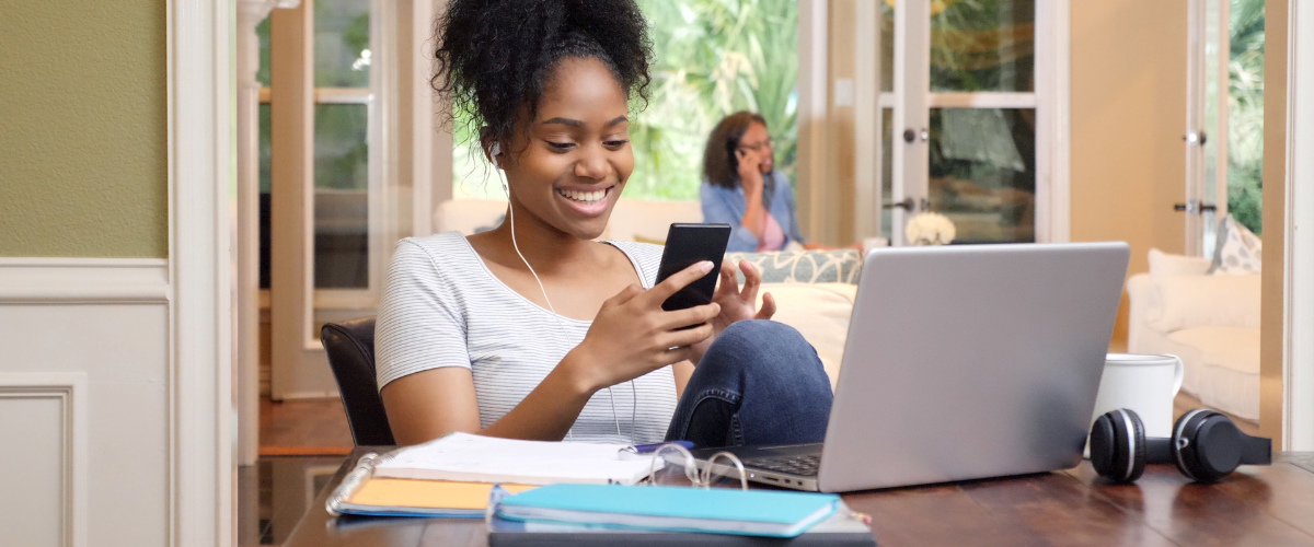 teen girl doing homework in front of laptop while also looking at phone