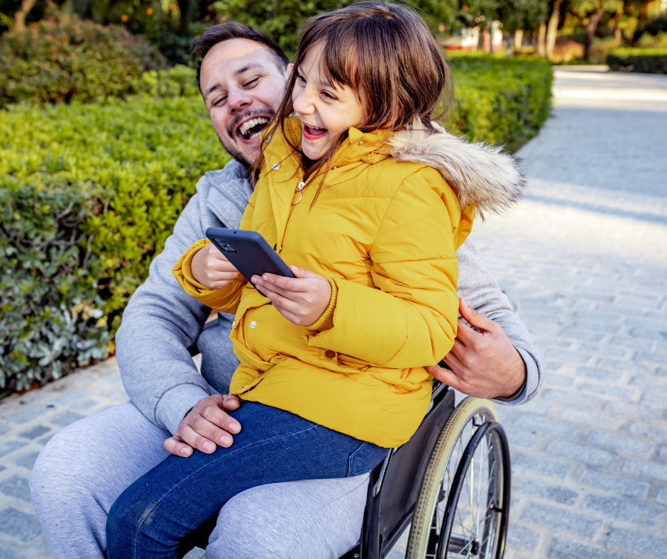 dad sitting in wheelchair with daughter on lap holding a phone, both laughing