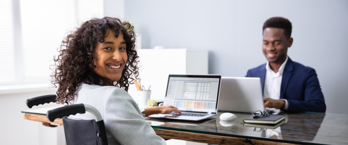 woman in wheelchair working at desk with laptop. man sitting across desk