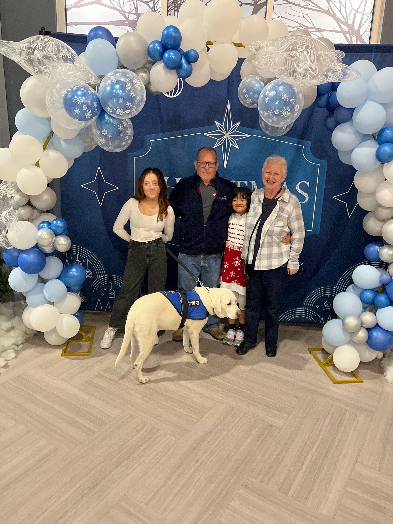 Tom with Marjorie, their two daughters, and Norma, standing and smiling for a family Christmas picture. Background of blue and white balloons surrounding a blue backdrop with star illustrations. Bare trees visible outside the high window behind.