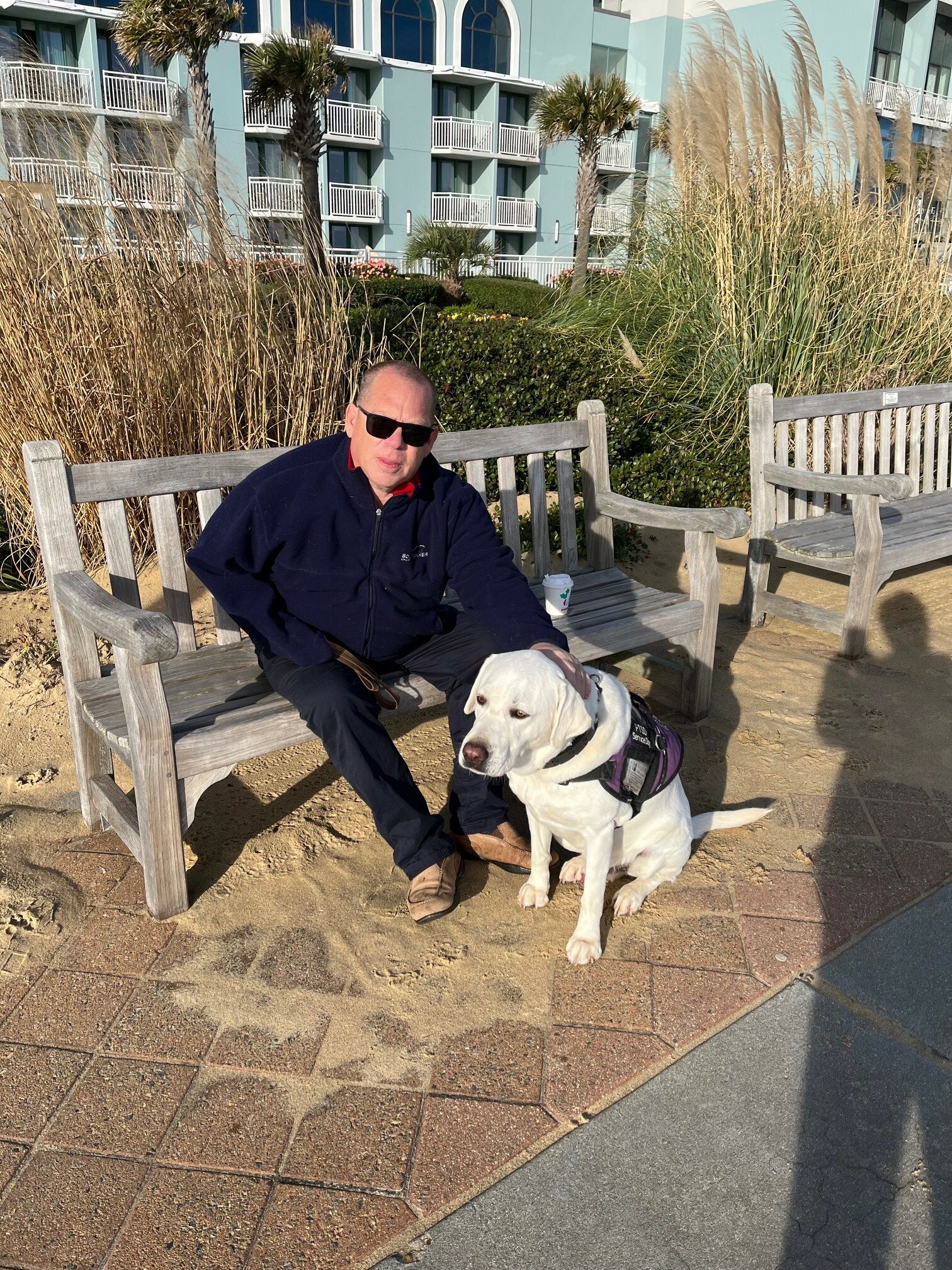 Tom Reichard (wearing sunglasses and a navy sweatshirt) on outdoor bench. Service dog (Norma), white retriever-type, sitting on ground next to him. Brick-paved walkway in front; in the background, tall brush and then a blue-green building with balconies, and two tall palm trees.