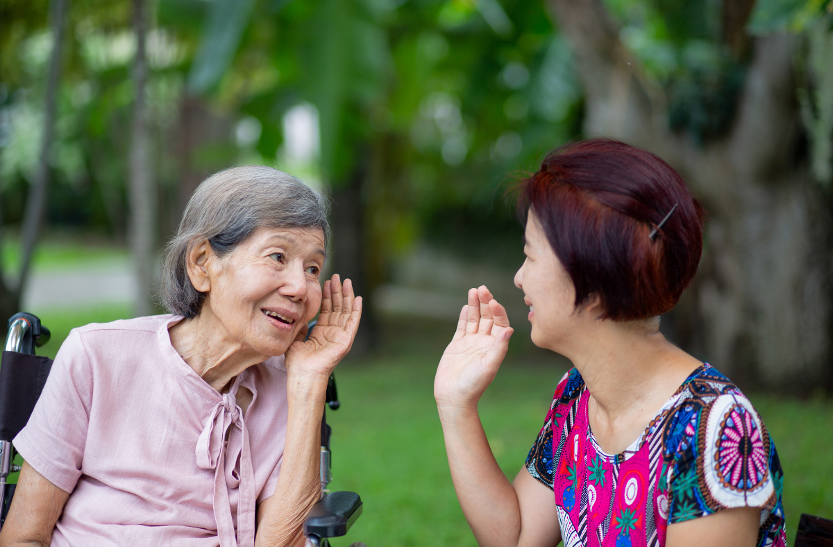 Two individuals with gray hair conversing outdoors; one seated in a wheelchair, the other closeby, gesturing as if sharing a story. Lush greenery forms the background.