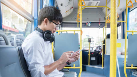 A young man wearing headphones and glasses sits on a bus, looking at his smartphone. The bus is relatively empty, and sunlight is streaming in through the windows.
