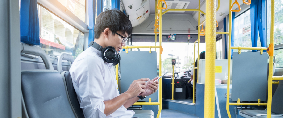 A young man wearing headphones and glasses sits on a bus, looking at his smartphone. The bus is relatively empty, and sunlight is streaming in through the windows.