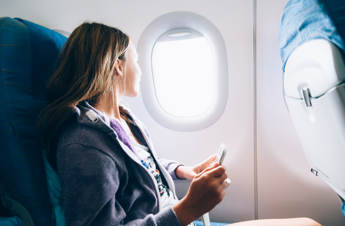 A young woman with long hair, wearing a casual hoodie, sits in an airplane seat by the window. She holds a smartphone in one hand and looks out the window, which is brightly lit by sunlight.