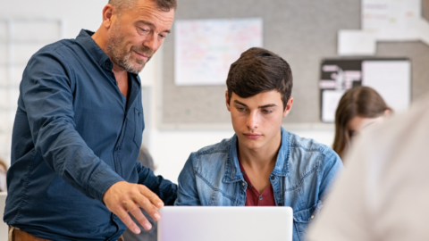 high school teacher helping young man with computer in classroom