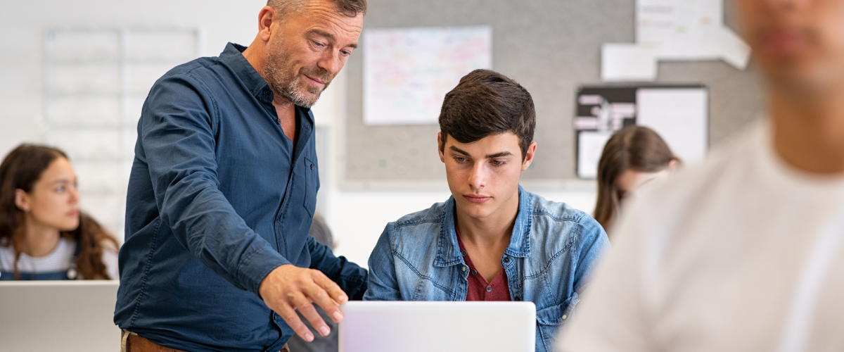 high school teacher helping young man with computer in classroom