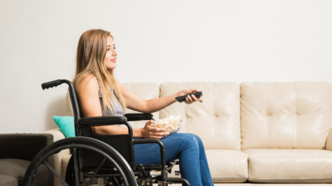 young adult woman sitting in wheelchair with bowl of popcorn on lap with tv remote in hand