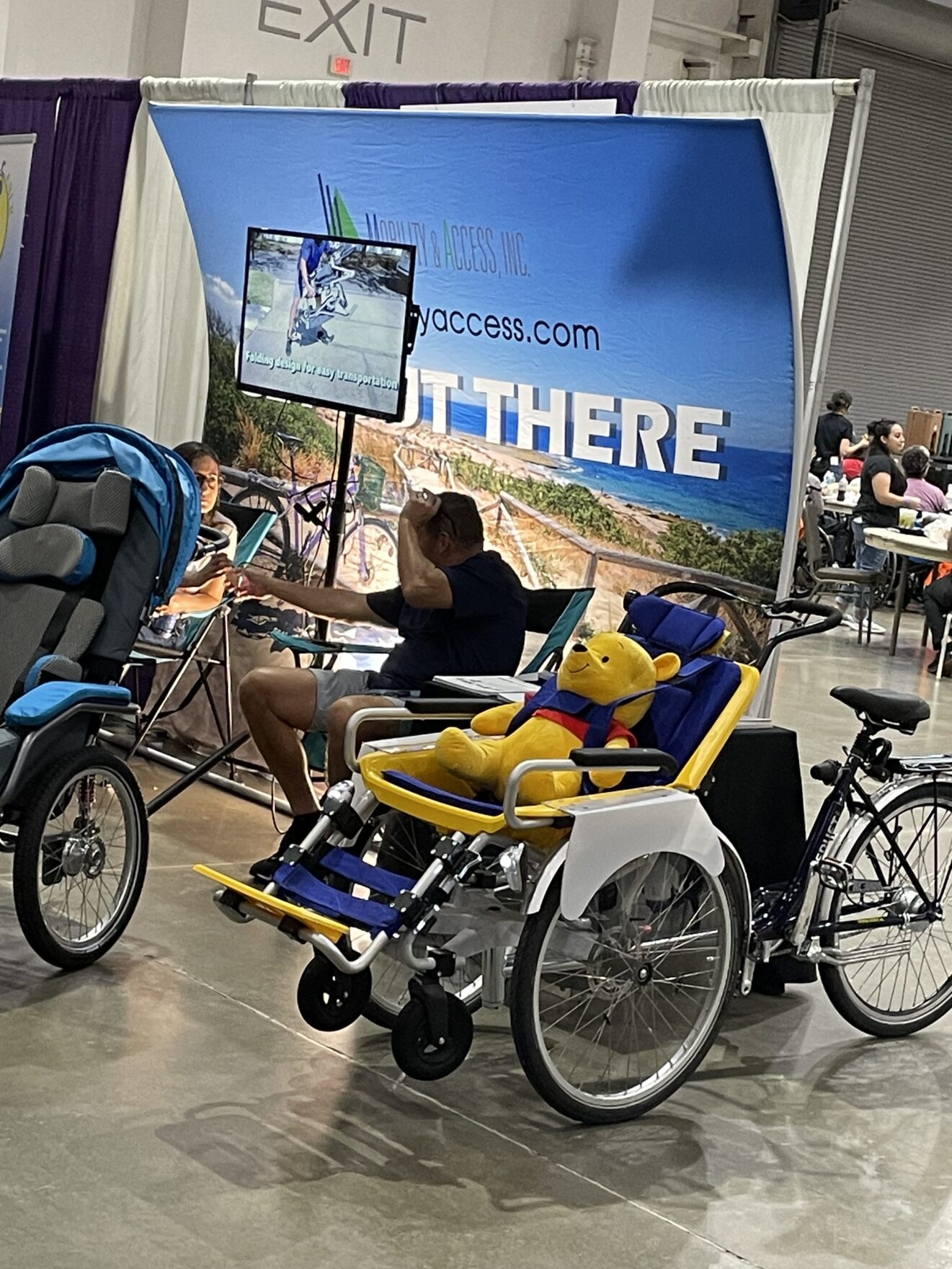 Mobility & Access Inc. booth. Display of travel wheelchairs and accessible bikes, with Winnie-the-Pooh teddy bear occupying seat in foreground.