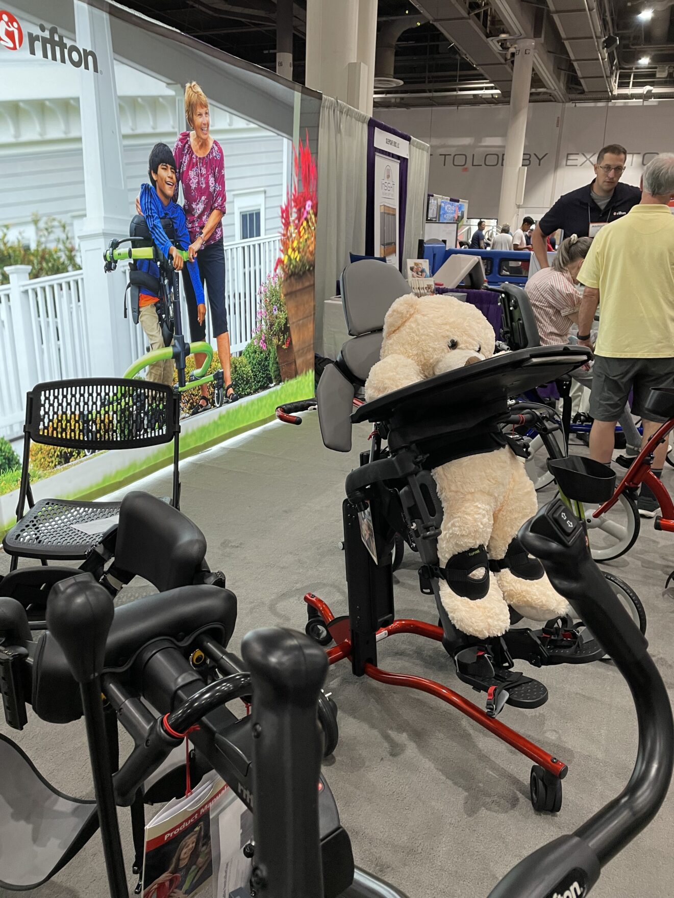 Rifton booth. Display of adaptive activity chairs, with large beige teddy bear in second chair in the foreground.