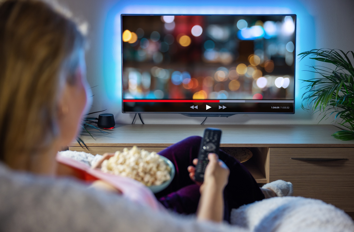 Person holding a remote and bowl of popcorn while watching a nighttime cityscape scene on a television in a cozy living room setting.