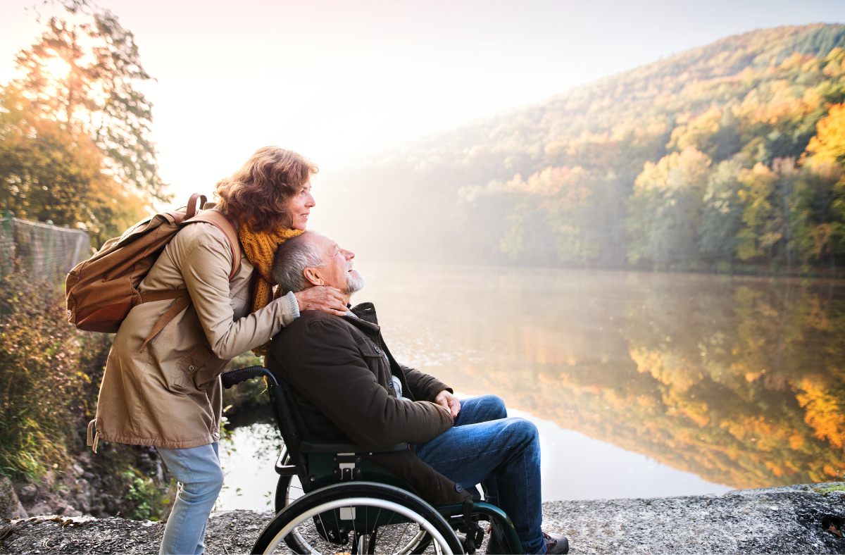 woman standing behind older man sitting in wheelchair. Couple is outside admiring fall foilage