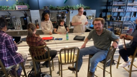 Group of people sitting and conversing around tables in a café with a "We see other people" sign in the background.