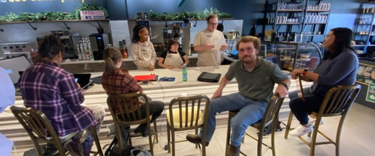 Group of people sitting and conversing around tables in a café with a "We see other people" sign in the background.