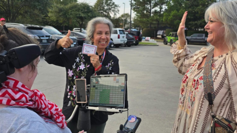 Three people in a parking lot celebrating a shared moment; one uses a wheelchair and speech-generating device, another shows a badge, and the third gestures a thumbs-up.