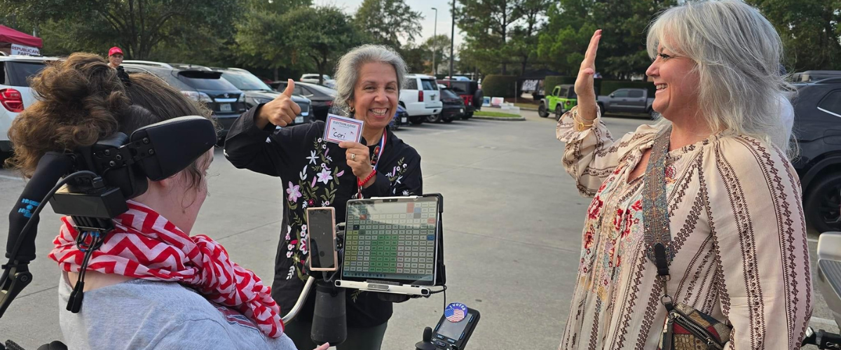 Three people in a parking lot celebrating a shared moment; one uses a wheelchair and speech-generating device, another shows a badge, and the third gestures a thumbs-up.