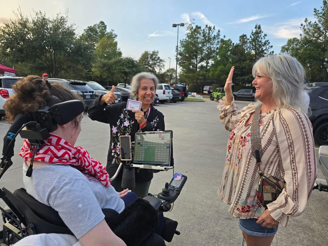 Three people in a parking lot celebrating a shared moment; one uses a wheelchair and speech-generating device, another shows a badge, and the third gestures a thumbs-up.