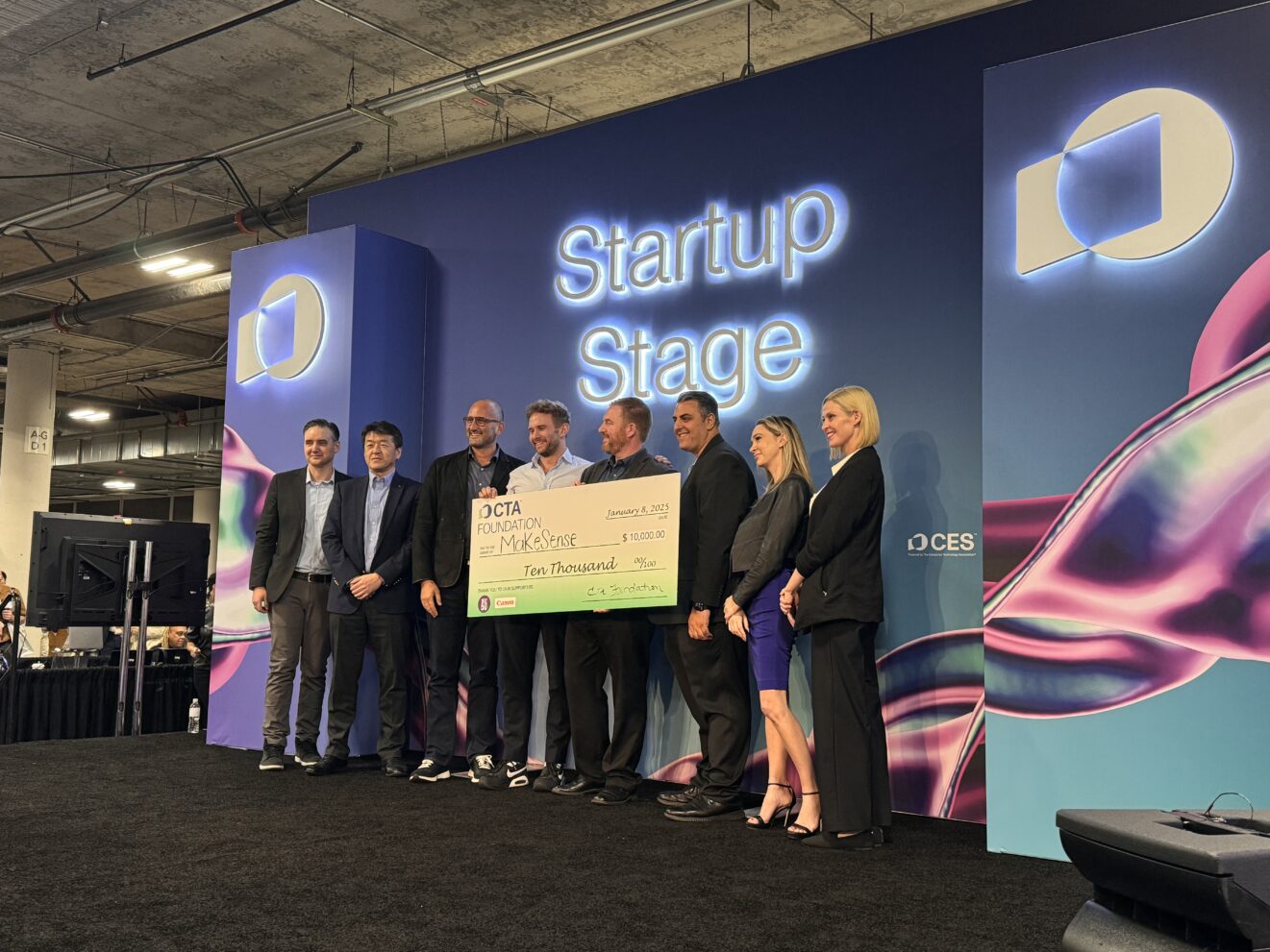 group of 8 people on stage under sign that reads 'startup stage', one person in middle holding a large check