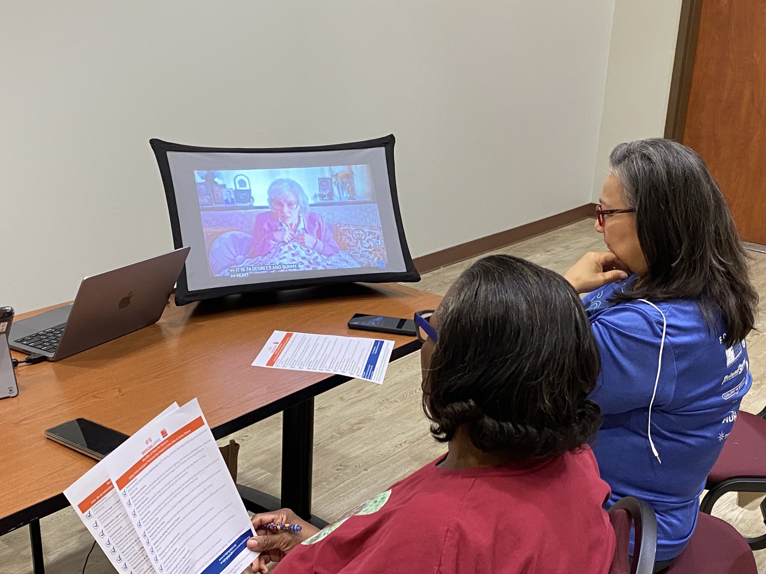 "Two women are seated in front of a projector screen in a classroom or workshop setting. They appear engaged and focused as they watch a presentation on technology, with the projector screen displaying text or images related to the lesson.