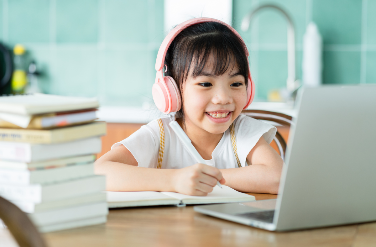girl with pink headphones writing in a notebook while looking at a laptop with a stack of books sitting nearby