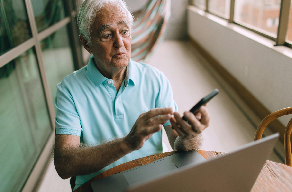 older adult man looking at phone while sitting in front of laptop