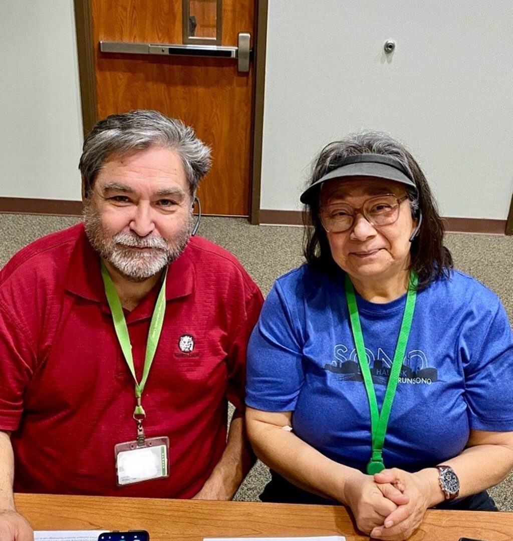 Fifty- or sixty-something couple sitting at Next50 table, wearing name tags hanging from green neck ribbons. Man, on left, is wearing a red shirt and has a short gray beard and a full head of mixed gray-and-black hair. Woman, on right, has mostly dark shoulder-length hair and is wearing glasses, a blue shirt, and a gray visor.