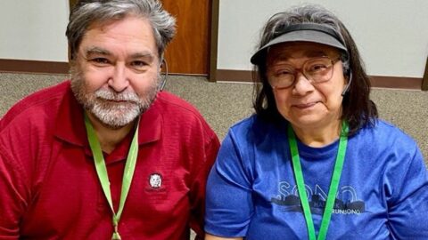 Fifty- or sixty-something couple sitting at Next50 table, wearing name tags hanging from green neck ribbons. Man, on left, is wearing a red shirt and has a short gray beard and a full head of mixed gray-and-black hair. Woman, on right, has mostly dark shoulder-length hair and is wearing glasses, a blue shirt, and a gray visor.