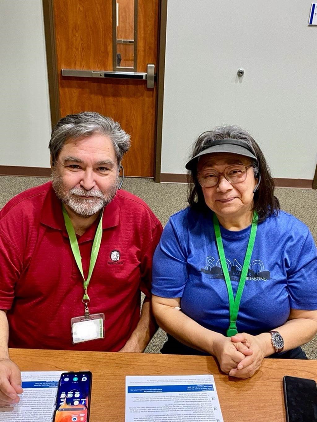 Fifty- or sixty-something couple sitting at Next50 table, wearing name tags hanging from green neck ribbons. Man, on left, is wearing a red shirt and has a short gray beard and a full head of mixed gray-and-black hair. Woman, on right, has mostly dark shoulder-length hair and is wearing glasses, a blue shirt, and a gray visor.
