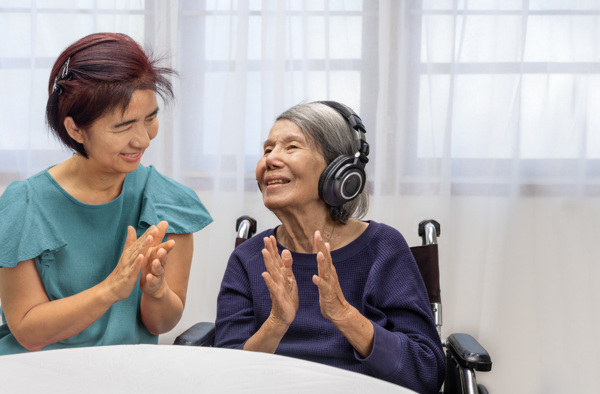 older woman in wheelchair wearing headphones smiling and clappying with friend