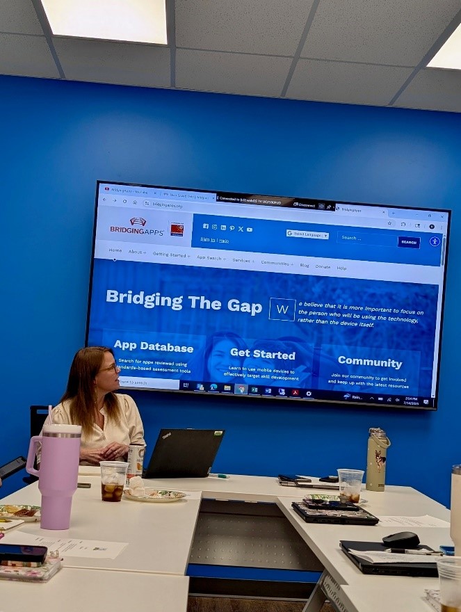Rectangle of business-lunch tables with open laptop computer at the head table. Worker seated in front of computer, turning head to the left to view overhead screen showing blue staging.bridgingapps.org home page.