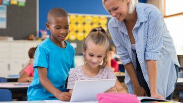 Ethnically diverse classroom. In foreground: girl with ponytail working on a laptop, with fellow student looking over her right shoulder, and adult looking over the left.