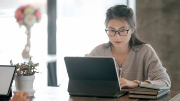 Teenage female student facing camera, looking down at laptop. Student has long, straight, dark hair and is wearing glasses and a beige V-neck sweater. Another laptop screen is partly visible in lower left corner, facing toward camera, with only a right hand showing for its user.