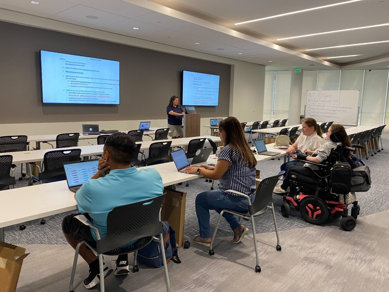 Classroom with rows of long tables, facing two blue screens of text. Whiteboard with writing visible to the right. Instructor in dark shirt stands at a podium between the screens. Line of four students, one in black-and-red wheelchair, seated at table in the foreground.