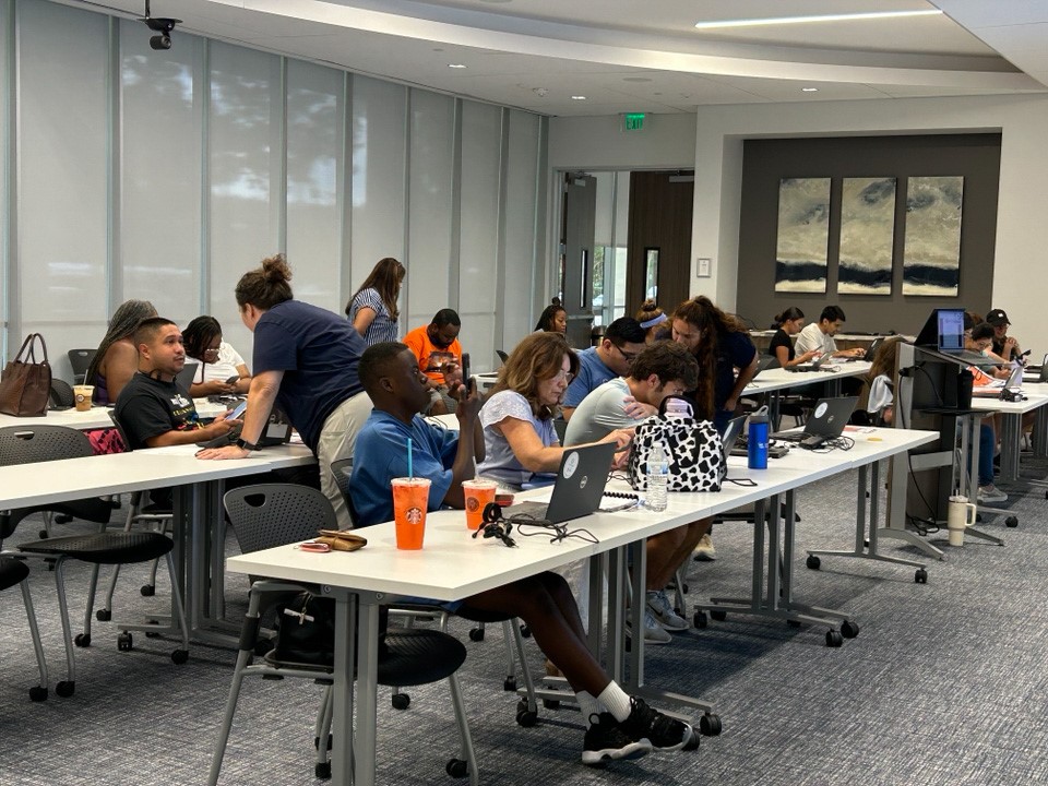 Front view of about a dozen students sitting at long tables. Three standing instructors are milling around. Four drink containers, one polka-dot handbag, and two open laptops visible beside students at front table.