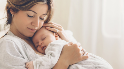 Mother gently holding and cuddling her sleeping baby while sitting on a bed in a softly lit room.