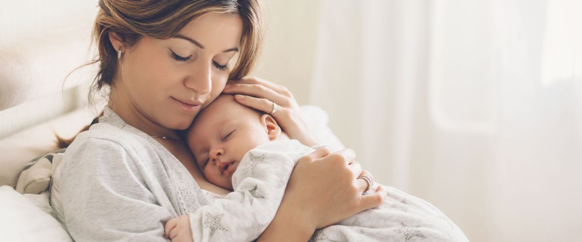 Mother gently holding and cuddling her sleeping baby while sitting on a bed in a softly lit room.