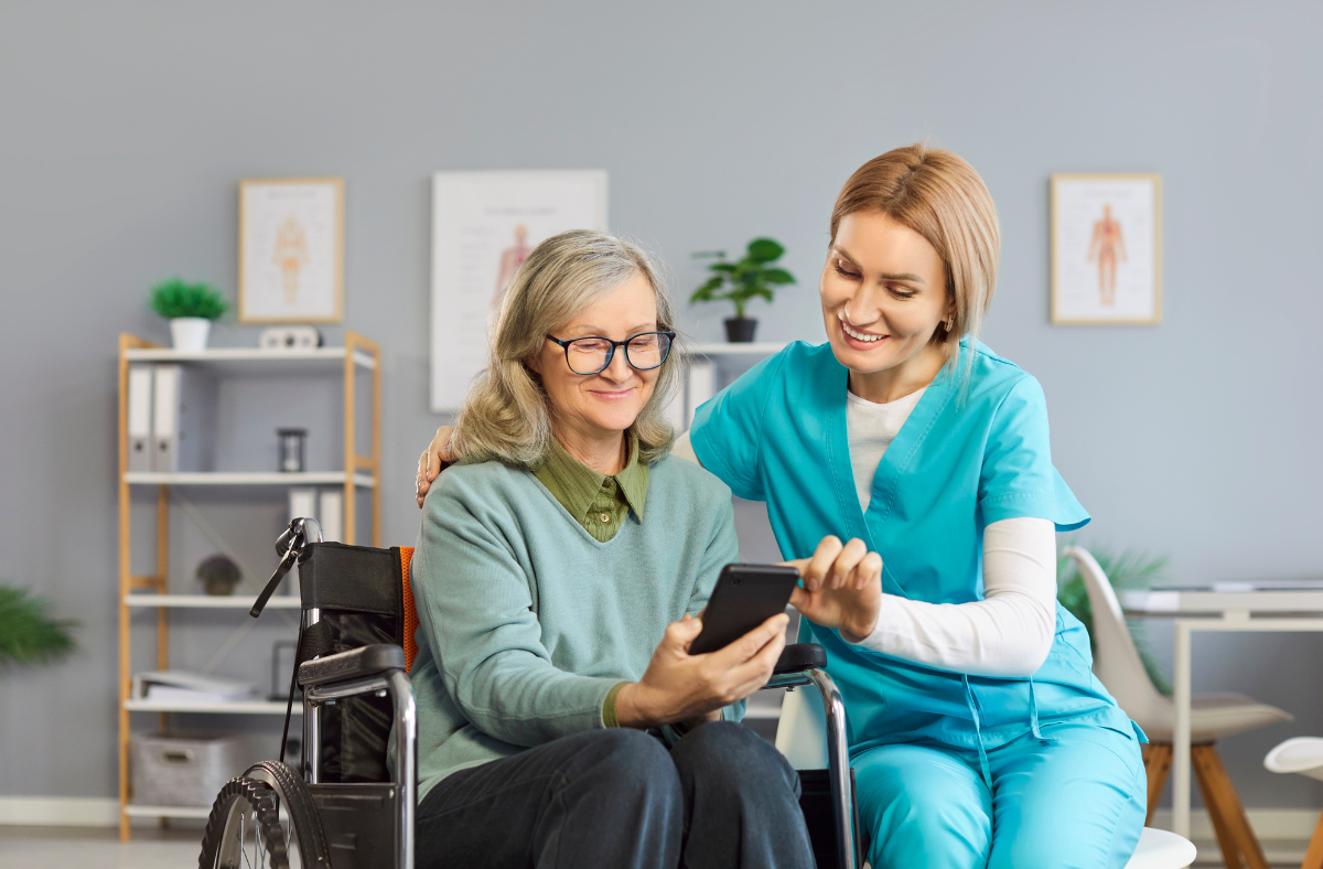 A nurse in blue scrubs sits beside an older woman in a wheelchair, smiling and showing her something on a smartphone in a bright medical office.