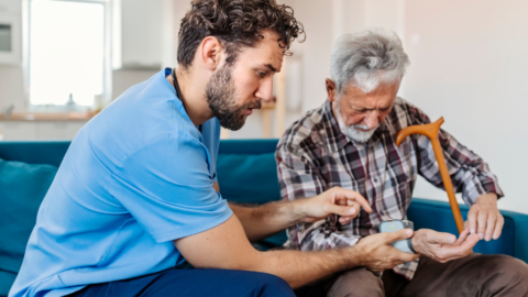 A healthcare worker helps an elderly man sitting on a couch check his blood sugar with a glucometer, while the man holds a wooden cane.