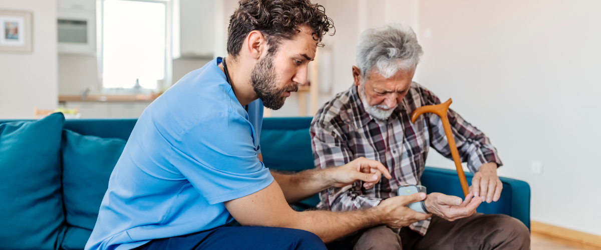 A healthcare worker helps an elderly man sitting on a couch check his blood sugar with a glucometer, while the man holds a wooden cane.