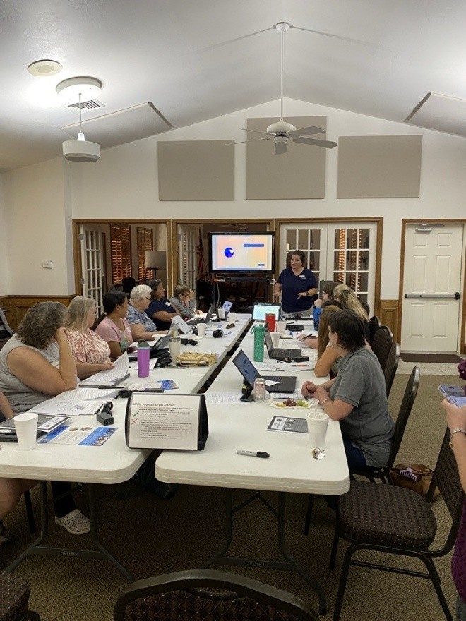 Group of people seated at long, white tables. Most have laptops in front of them. At the head of the tables, a trainer stands to the right of a video screen showing a blue circle and blue bar.